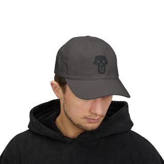 A man wearing a black hoodie and a dark cap featuring a skull logo, styled in casual men’s graphic streetwear.
