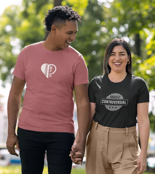 A couple holds hands, wearing matching graphic t-shirts in a sunny outdoor setting, surrounded by green trees.
