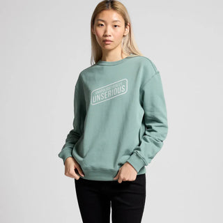 A women wearing a mint green sweatshirt with the word "UNSERIOUS" printed on the front, paired with black pants, standing against a plain backdrop.