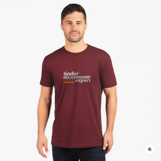 A man wears a maroon t-shirt featuring the text "tinder RELATIONSHIP expert" in a casual setting. 